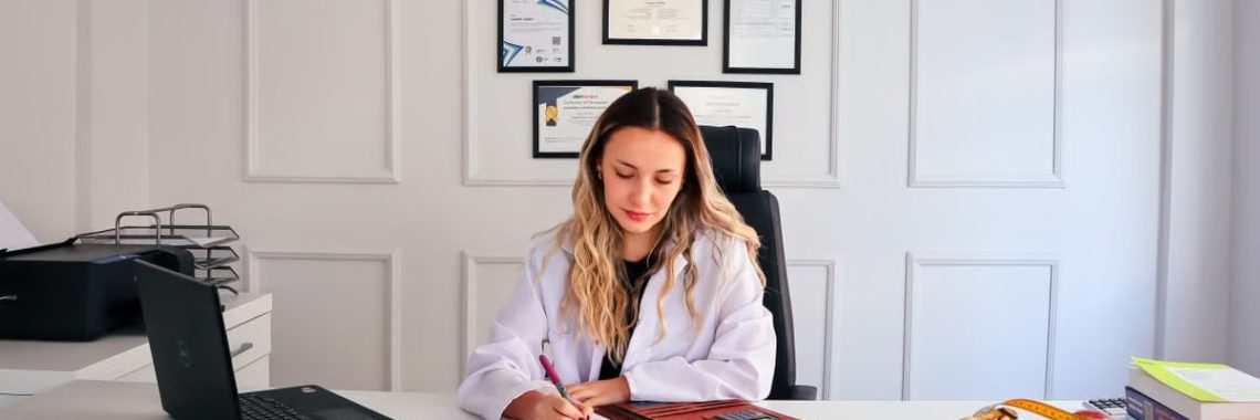 Nutritionist sits at a modern office desk writing, with fruits and diplomas displayed, emphasizing health and professionalism.