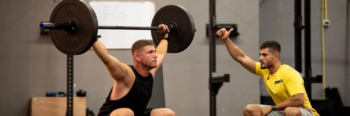 Athlete performing weightlifting exercise under trainer's guidance in a gym setting.